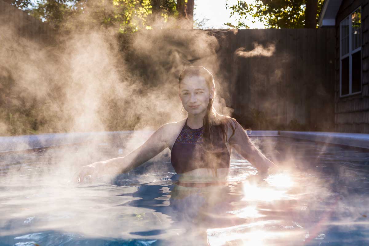 A young woman enjoys a hot tub outdoors – thick steam rises from the blue water, surrounded by rays of sunshine and a wooden fence, creating a cozy sauna and spa atmosphere