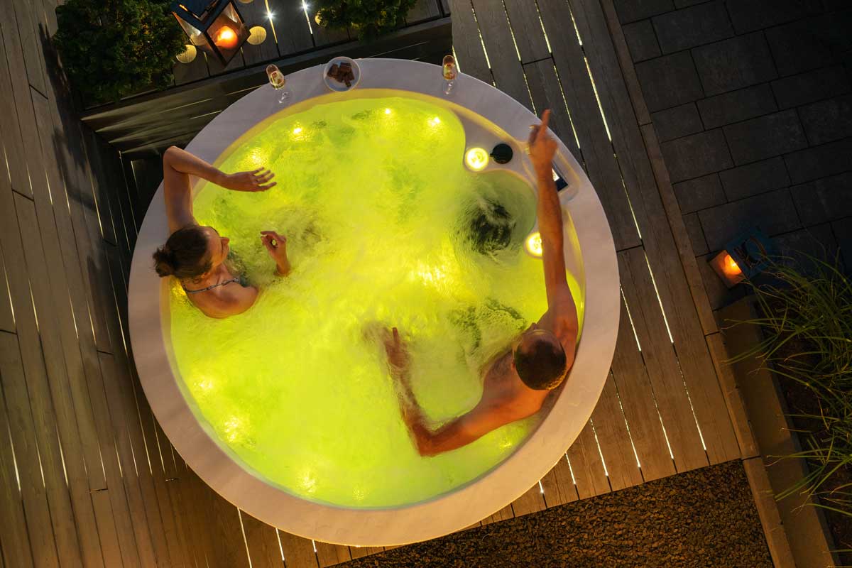 Two people are relaxing in a round outdoor jacuzzi tub with bright yellow water lighting, bubbling bubbles, decorative plants and lit candles all around, creating a romantic spa atmosphere