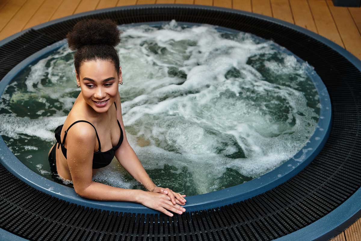 A woman in a black swimsuit smiles as she sits on the edge of a round bubble jacuzzi. The water bubbles, and the modern wooden floor adds coziness to the contemporary spa environment with a hot tub