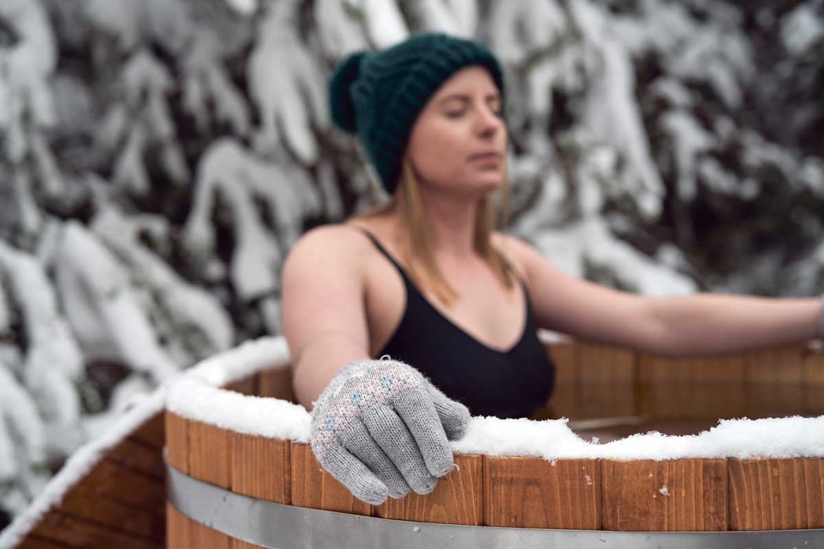 A woman wearing a knitted green hat and gray woolen gloves enjoys a hot outdoor hot tub in winter, the woodwork covered with a blanket of white snow, and snow-covered trees in the background – a contrast between cold and warmth