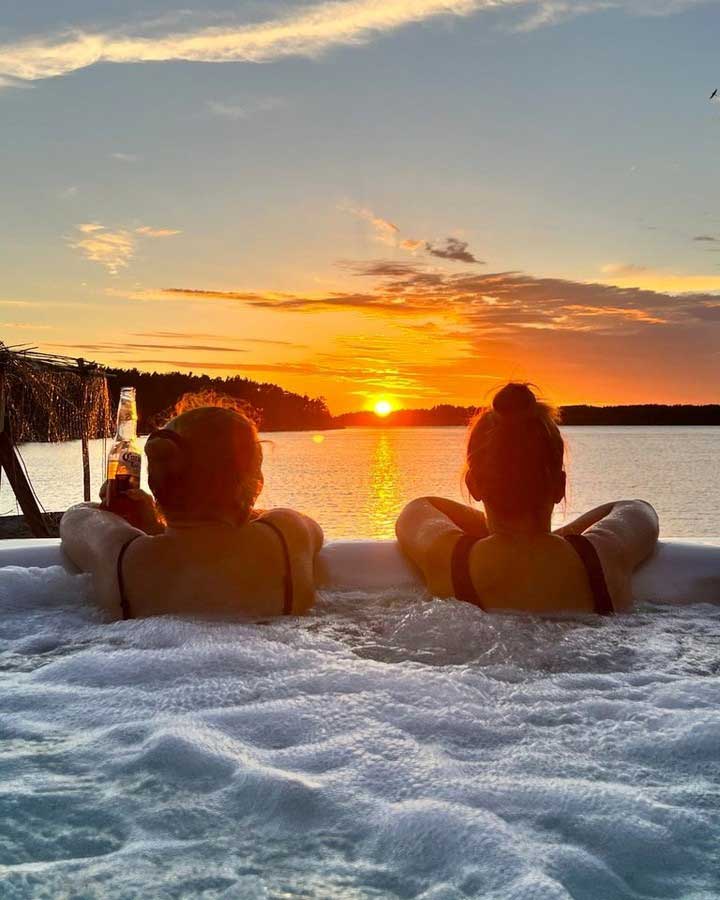 Two women are sitting in a white outdoor jacuzzi, enjoying the bubbles and watching the spectacular orange sunset over the lake, one holding a bottle of drink in her hand – a party moment by the water