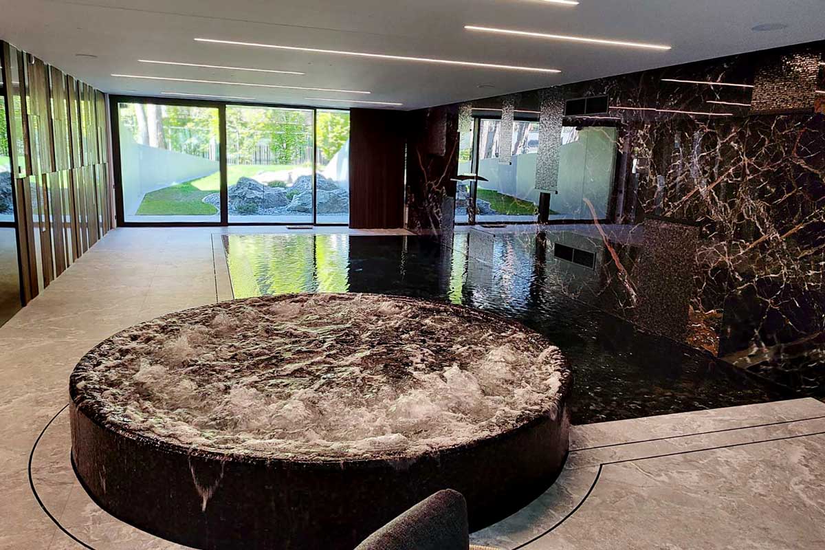 Luxurious SPA interior with a round black marble jacuzzi, bubbling water, and a large swimming pool nearby, shiny stone walls, and wide glass doors opening onto a green garden – a modern relaxation area