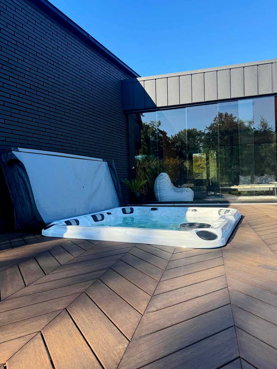 A white outdoor jacuzzi is set on a modern wooden terrace next to the house with a black brick wall. Clear water flows in the tub, and the glass walls of the house and blue sky provide the backdrop for a cozy spa corner in the yard