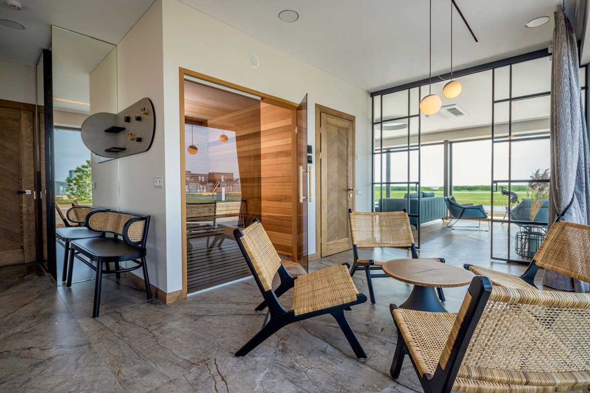 A modern SPA relaxation area separated by a glass wall from a sauna with wooden walls and benches, with wicker chairs and a round table in front, and a living room with panoramic windows overlooking the countryside in the distance