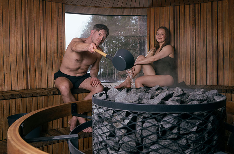A man pours water onto the heated sauna stones with a ladle, a woman sits nearby; a steamy, round wooden sauna with a window.