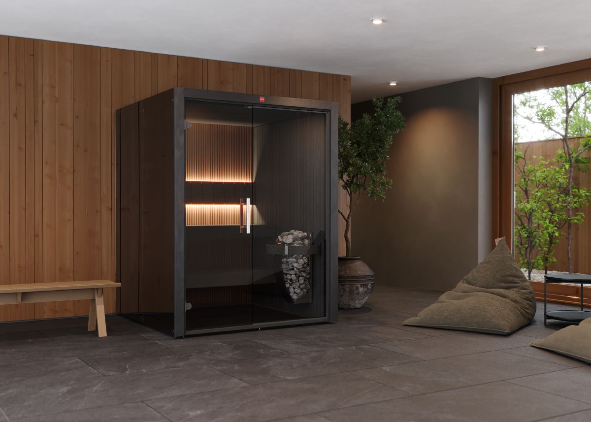 A modern glass sauna with black frames and interior lighting, built in a spacious room with wooden walls, decorative plants, and a relaxation area.