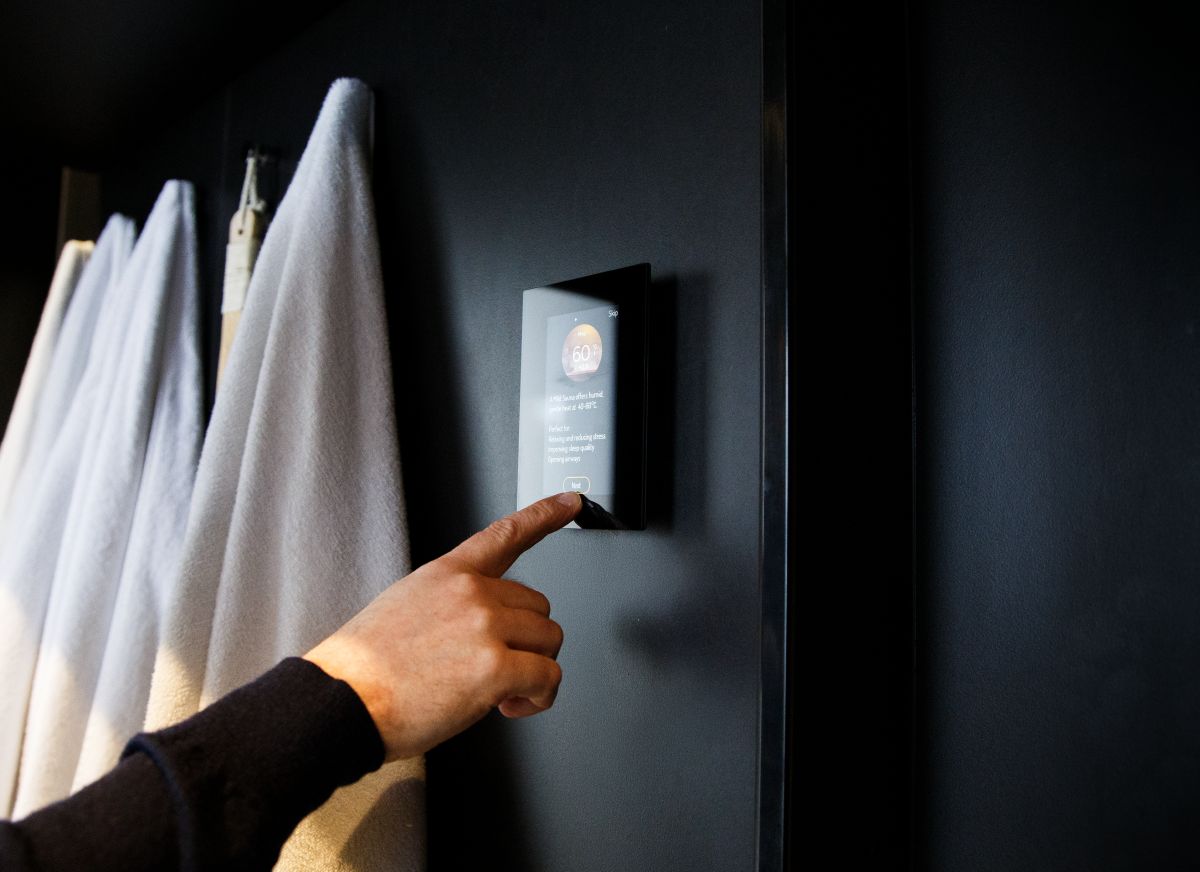 A man presses a modern wall-mounted sauna control panel with a digital display, white towels hanging nearby.