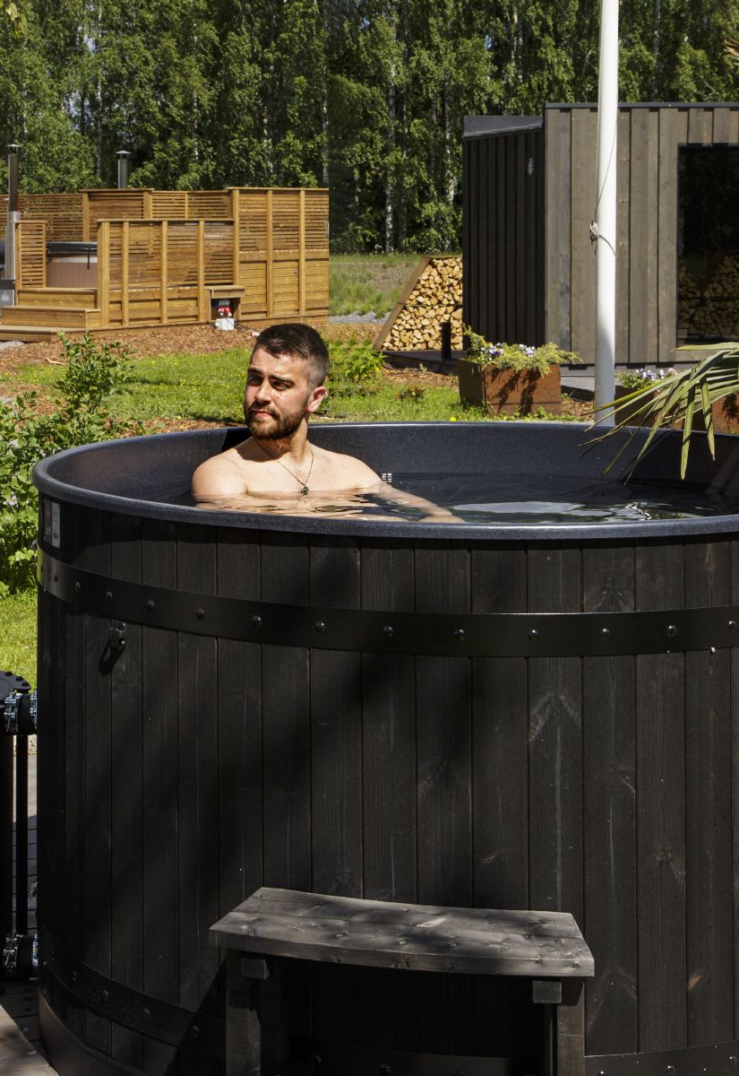 A man is sitting in an outdoor hot tub in a garden surrounded by wooden saunas, piles of firewood, and greenery
