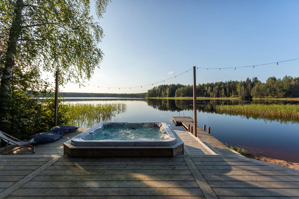 Outdoor jacuzzi on a wooden terrace by the lake, with a pier, surrounded by nature and fairy light lighting