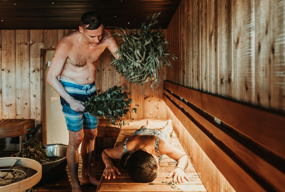 Vīrietis pirtī sit sievieti ar bērza slotiņu — tradicionāls pirts rituāls atpūtai.
